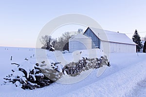 Low angle view of piles of cut wood and farm buildings covered in fresh snow during a blue hour winter morning