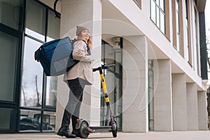 Low-angle view of positive female courier with big thermo backpack standing with electric scooter in city street looking