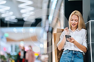 Low-angle view of pretty blonde young woman with long hair in stylish clothing using mobile phone, holding shopping