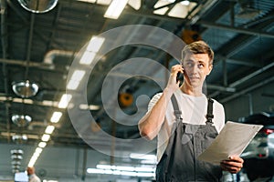 Low-angle view of professional handsome young mechanic male wearing uniform holding clipboard and talking on mobile