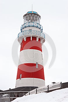 A low angle view of a red and white striped lighthouse with white wooden fence leading to it. A house is on the bottom of the