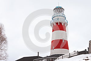 A low angle view of a red and white striped lighthouse with white wooden fence leading to it. A house is on the bottom of the