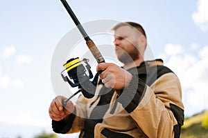 Low-angle view selective focus of fisherman holding casting rod wearing raincoat standing on bank waiting for bites on