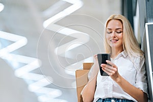 Low-angle view of smiling attractive blonde young woman with long hair in stylish clothing using mobile phone, holding