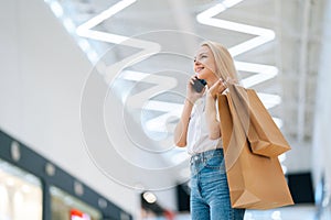 Low-angle view of smiling attractive blonde young woman in stylish clothing talking on mobile phone, holding shopping
