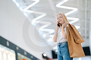 Low-angle view of smiling attractive blonde young woman talking on mobile phone looking at camera, holding shopping