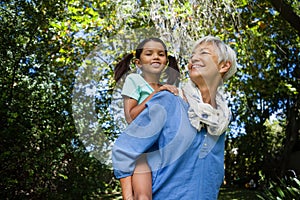 Low angle view of smiling grandmother giving piggyback to granddaughter against trees