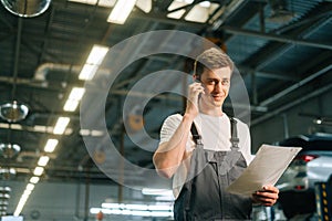 Low-angle view of smiling handsome young mechanic male wearing uniform holding clipboard and talking on mobile phone.