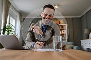 Low angle view of smiling male manager holding glasses and signing document on desk at home.