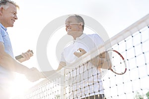 Low angle view of smiling men shaking hands while standing at tennis court against clear sky