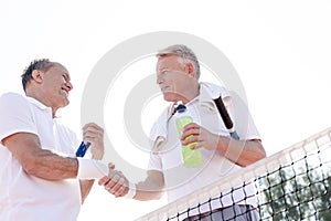 Low angle view of smiling men shaking hands while standing at tennis court against clear sky on sunny day