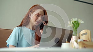 Low-angle view of smiling young redhead woman talking on smartphone and using laptop sitting at table by window in cafe.