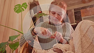 Low angle view of teenaged girl sitting in chair near window and knitting. Cosy evening at home.