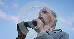 Low angle view of traveler looking into field-glass on blue sky background slow motion. Serious man exploring horizon in