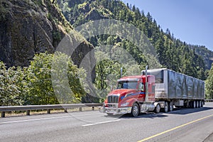 Low cab profile bright red big rig semi truck transporting cargo in covered framed semi trailer moving on the wide highway road
