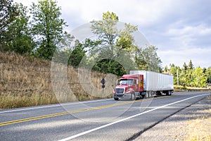 Low cab profile red big rig semi truck transporting goods in dry van semi trailer driving on the narrow mountain road