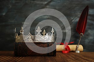 Low key image of beautiful queen/king crown, old books and feather quill ink pen over wooden table. fantasy medieval period