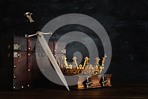 low key image of beautiful queen/king crown over antique book, chest and sword. fantasy medieval period.