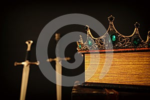 low key image of beautiful queen/king crown over antique book next to sword. fantasy medieval period. Selective focus