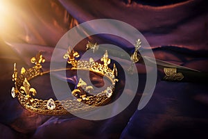 Low key image of beautiful queen/king crown and sword over dark royal purple delicate silk. fantasy medieval period