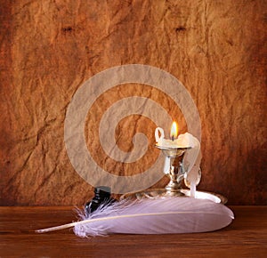 Low key image of white Feather, inkwell and burning candle on a wooden table.