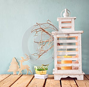 Low key image of white wooden vintage lantern with burning candle, wooden deer, christmas gifts and tree branches on wooden table.