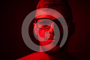 Low key portrait of bearded young man in glasses looking at camera in red light