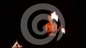 Low key. A young male with long hair and a naked torso rotates a burning torch outdoors on a black night video slow