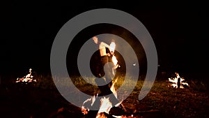 Low key. A young male with long hair and a naked torso rotates a burning torch outdoors on a black night video slow