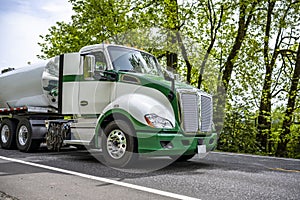 Low profile day cab big rig green and white semi truck transporting liquid cargo in tank semi trailer running on the narrow road