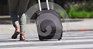 Low section of african american woman crossing road wheeling suitcase