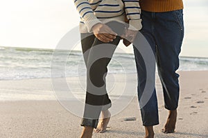 Low section of biracial couple holding hands while walking together on shore at beach during sunset