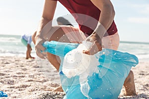 Low section of biracial man collecting plastic waste in bag at beach on sunny day