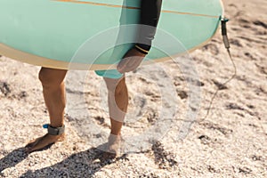 Low section of biracial senior man carrying surfboard standing barefoot on sand at sunny beach