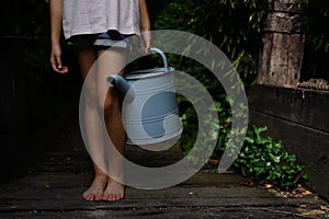 Low section of little girl in shorts holding watering can outdoors in garden.