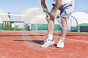 Low section of mature man holding tennis racket while suffering from knee pain on red tennis court during summer