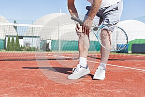 Low section of mature man holding tennis racket while suffering from knee pain on red tennis court during summer photo