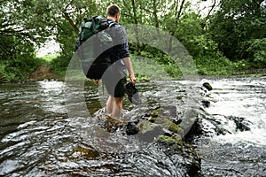 Low section shot of a man in a plaid shirt with heavy hiking backpack crossing the streem barefoot