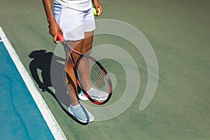 Low section of young female african american athlete standing with tennis racket and ball at court