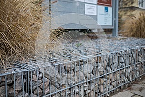 Low stylish gabion cage wall as barrier between street and house with reed grass and display sign in blurred background