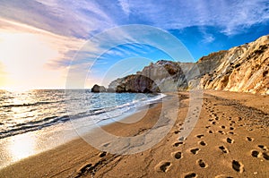 Low sun over beach, cliffs and hills and deep azure sea. Agios Ioannis beach, Milos island, Greece.