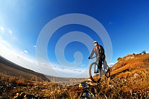 Low, wide angle portrait against blue sky of mountain biker going downhill. Cyclist in black sport equipment and helmet