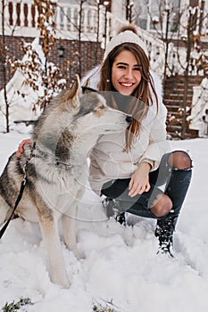 Loyal dog looking away in winter day while laughing girl in white jacket stroking him. Spectacular european lady in