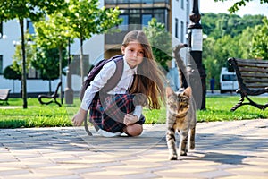 Lttle girl in uniform and with backpack playing with a stray cat on the street
