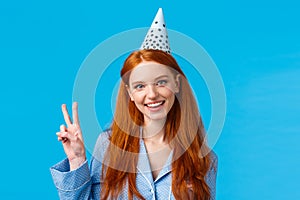 Lucky and enthusiastic feminine pretty redhead woman with long curly hair in nightwear, wearing birthday cap showing