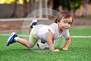 Lucky goalkeeper: little football player caught soccer ball, goalposts in background. Toddler boy lying on belly on soccer ball