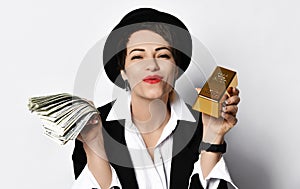 Lucky woman in stylish hat and black jacket is holding a fan of dollars and a bar of gold in her hands sends us a kiss