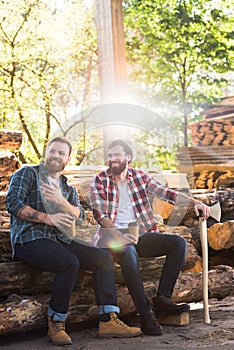 lumberjack with coffee cup pointing by finger to partner with axe sitting on logs