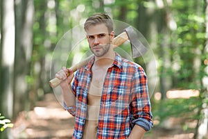 Lumbersexual guy in lumberjack shirt holding axe on shoulder forest background