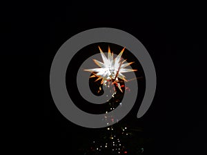 Luminous multi-pointed star at the top of a festively decorat-ed Christmas tree in an outdoor location in night, under the